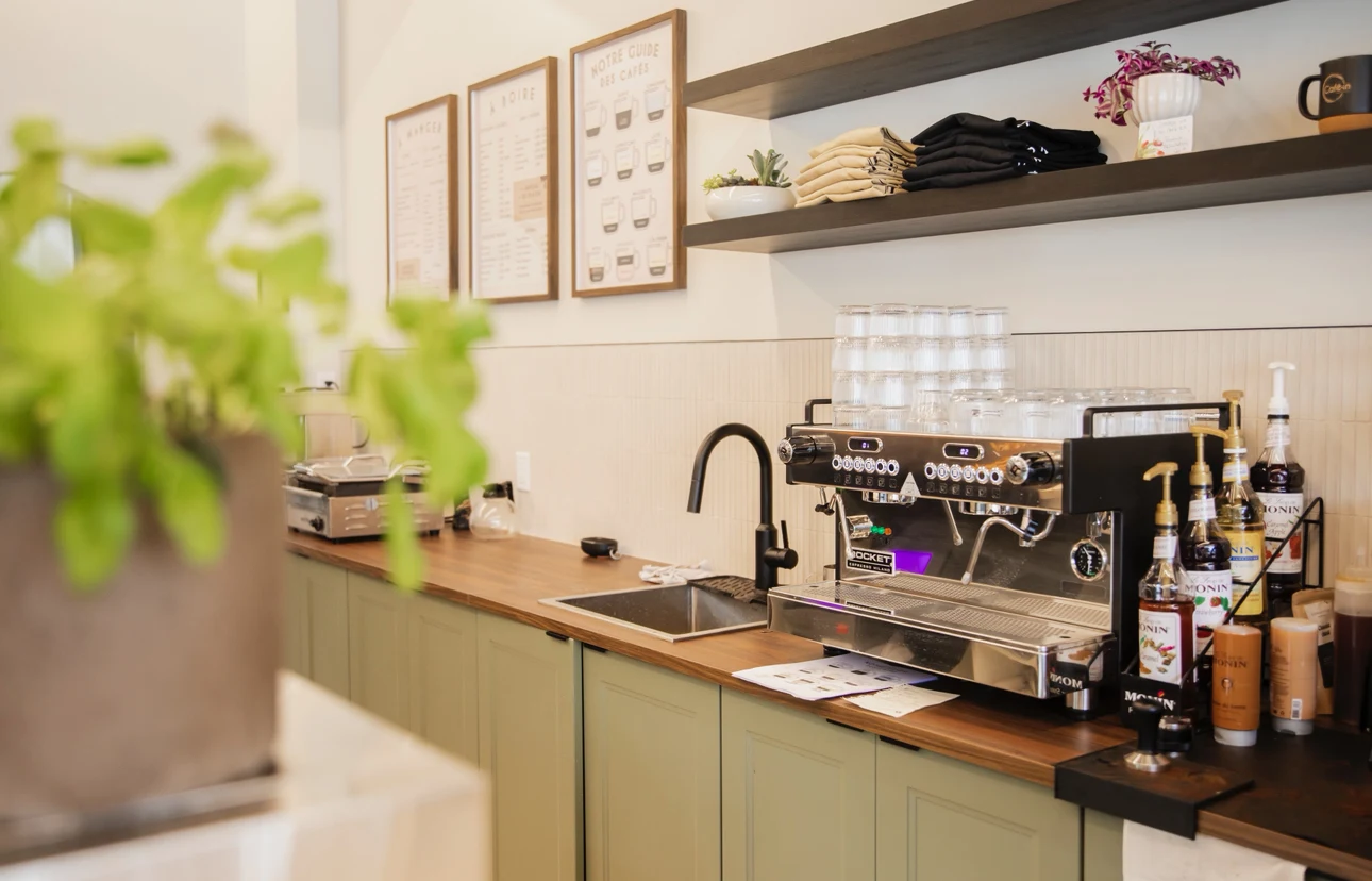 Le bar à café : machine espresso, armoires vert sauge, comptoir de bois et tableaux du menu au mur